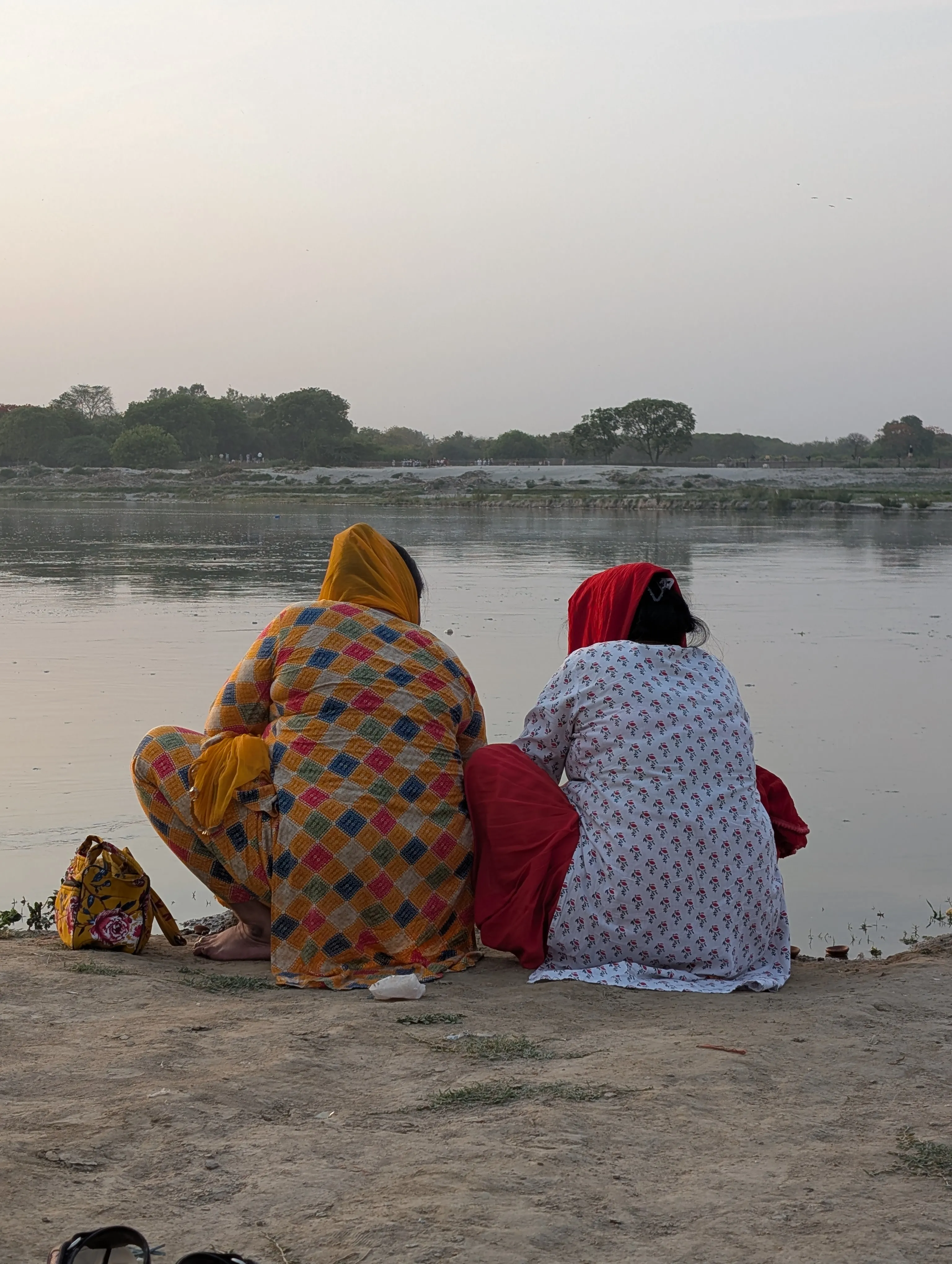Two women in kurtas sit by the river at dusk.